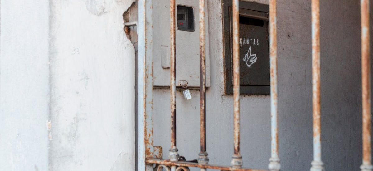 A rusty metal fence frames an old mail drop against a worn wall, creating a rustic atmosphere.