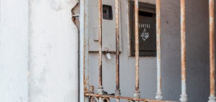 A rusty metal fence frames an old mail drop against a worn wall, creating a rustic atmosphere.