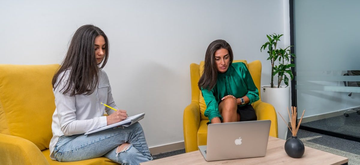 Two women work in a modern office setting, seated on yellow chairs, focusing on a laptop and notes.