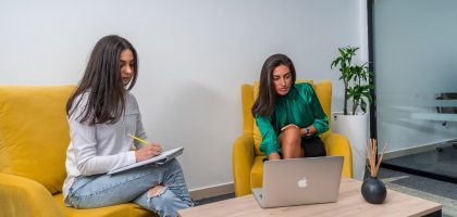 Two women work in a modern office setting, seated on yellow chairs, focusing on a laptop and notes.