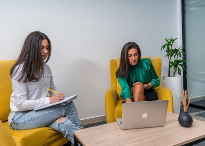 Two women work in a modern office setting, seated on yellow chairs, focusing on a laptop and notes.